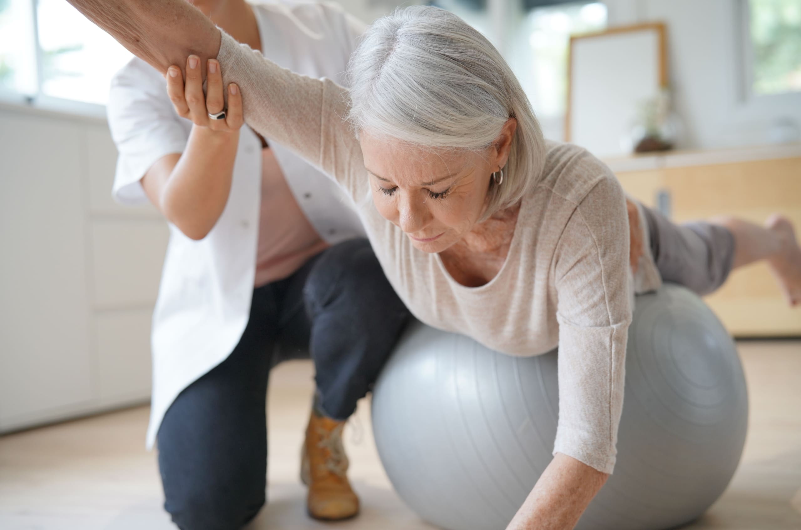 Physiotherapist helping an elderly patient use a balance ball