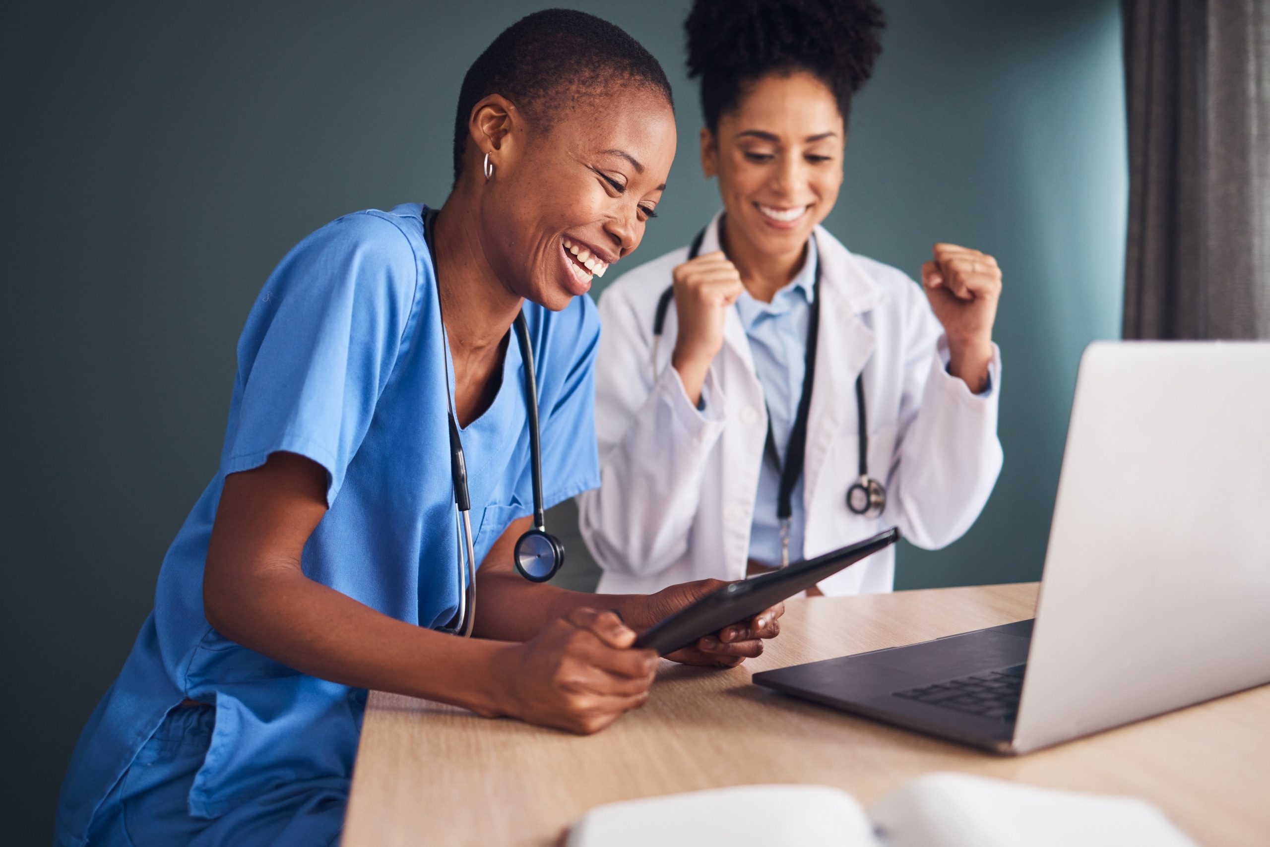 Two nurses laughing together as they use a tablet and laptop